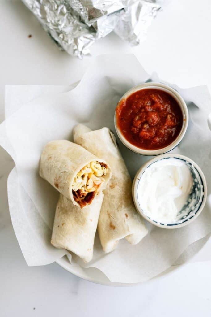 Breakfast burritos on a plate, served with bowls of salsa and sour cream. Wrapped burritos are cut in half, showing ingredients inside. Foil-wrapped item in the background.