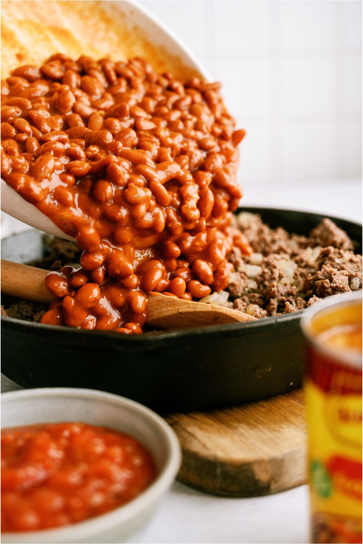 Adding beans into a skillet of cooked ground beef with wooden spoon.