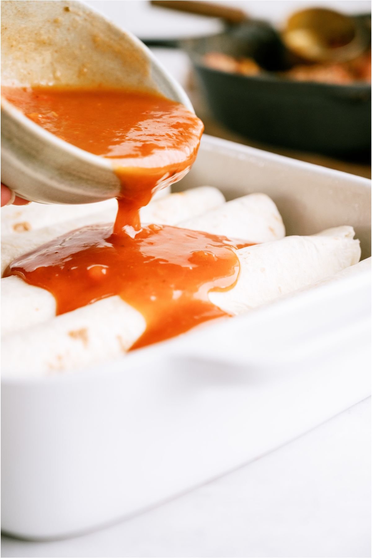 Pouring enchilada sauce over rolled enchiladas in baking dish.