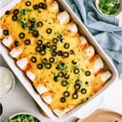 A baking dish filled with enchiladas topped with melted cheese and sliced black olives, surrounded by bowls of lettuce, sour cream, and chopped herbs.