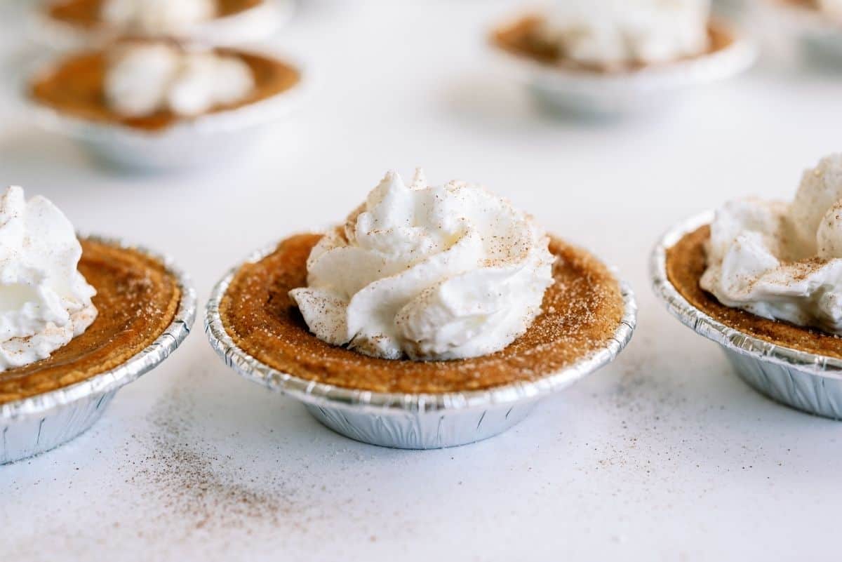 Mini pumpkin pies in foil tins topped with whipped cream and a sprinkle of cinnamon.