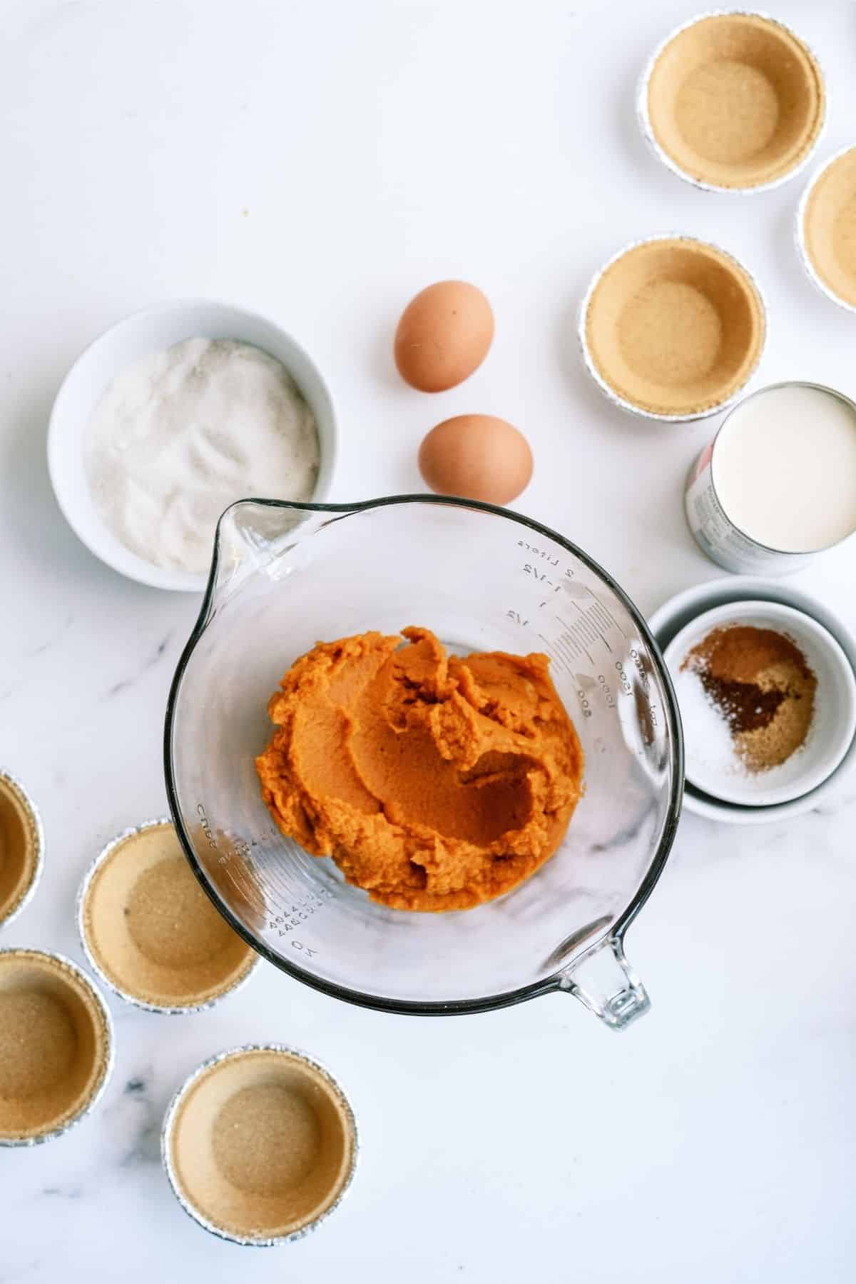 A bowl of pumpkin puree, two eggs, sugar, spices, and several premade pie crusts are arranged on a white surface, ready for making mini pumpkin pies.