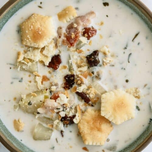 A bowl of creamy clam chowder topped with oyster crackers, chunks of potato, and pieces of bacon.
