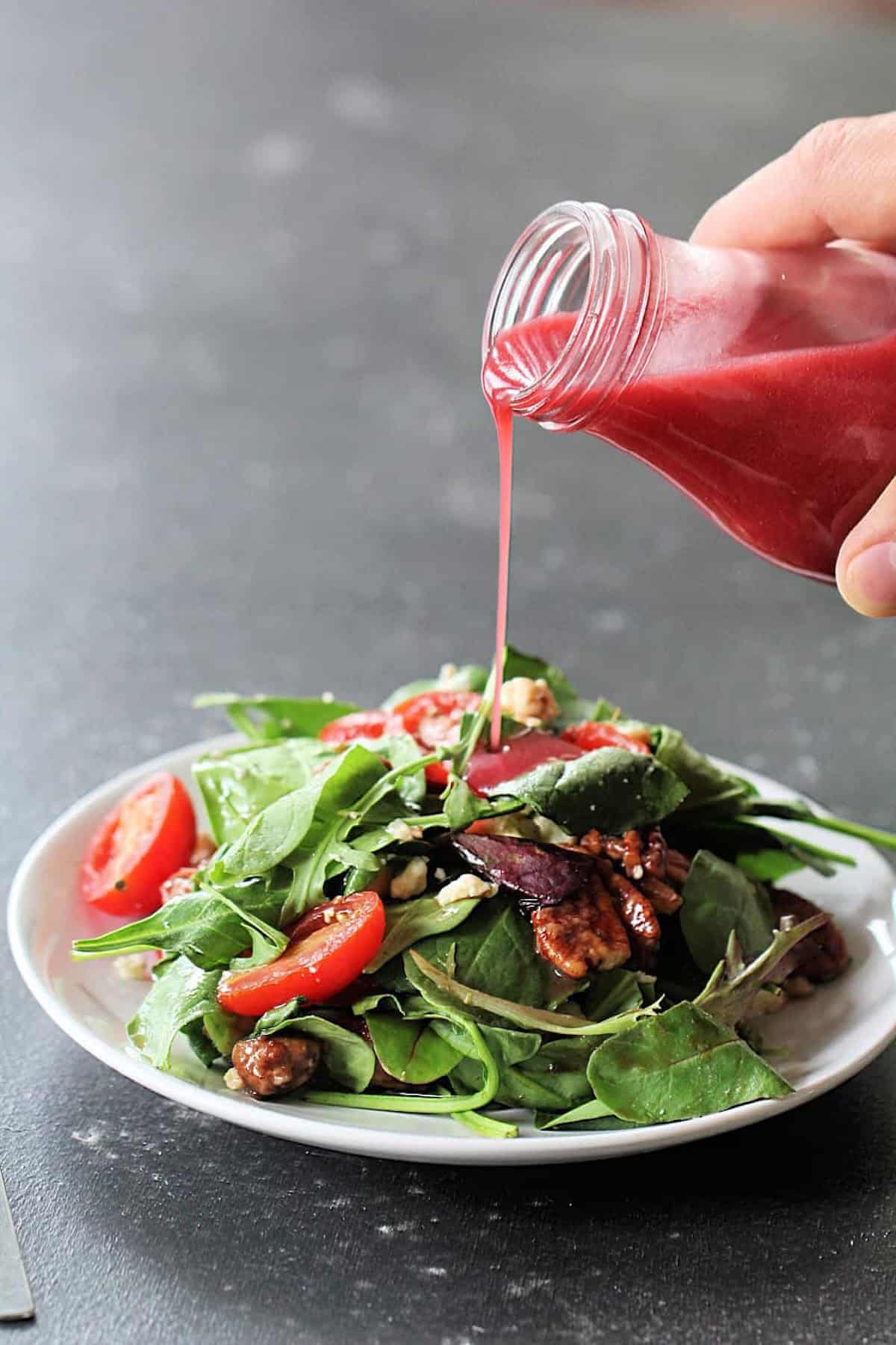 A hand pours red salad dressing over a plate of mixed greens, tomatoes, walnuts, and cheese.
