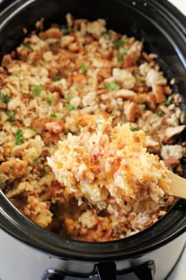 A close-up of a spoon scooping a serving of rice and vegetable dish from a black pot. The dish appears to include rice, chopped vegetables, and herbs.