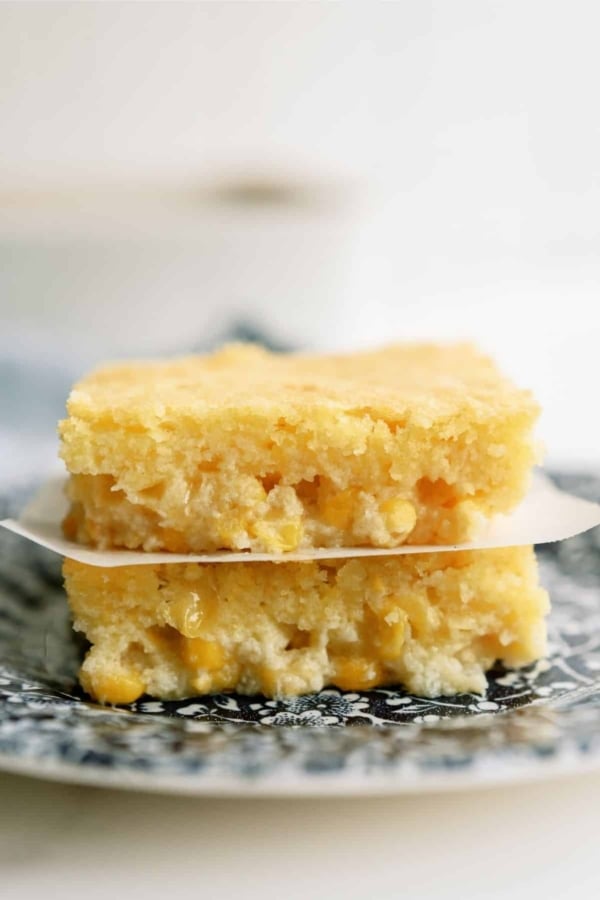 Two pieces of cornbread casserole are stacked on a plate. The cornbread has a golden yellow color and visible corn kernels.