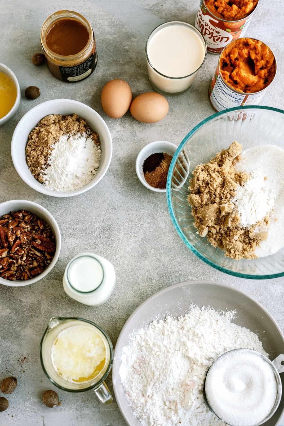 Ingredients for baking laid out on a table: flour, brown sugar, white sugar, pecans, eggs, butter, milk, spices, canned pumpkin, and a glass of milk.