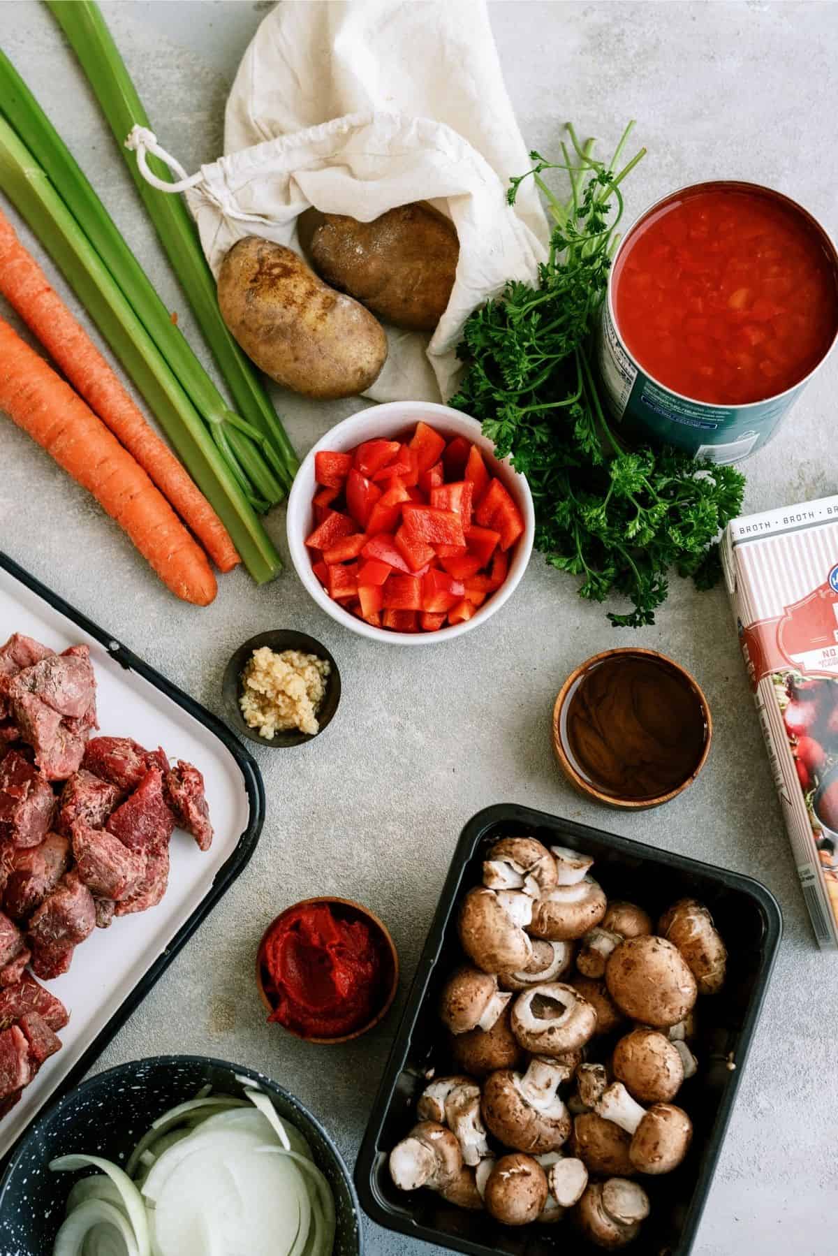 A variety of raw ingredients on a countertop: celery, carrots, potato, parsley, canned tomatoes, chopped red bell pepper, minced garlic, uncooked beef, tomato paste, mushrooms, onions, and beef broth.