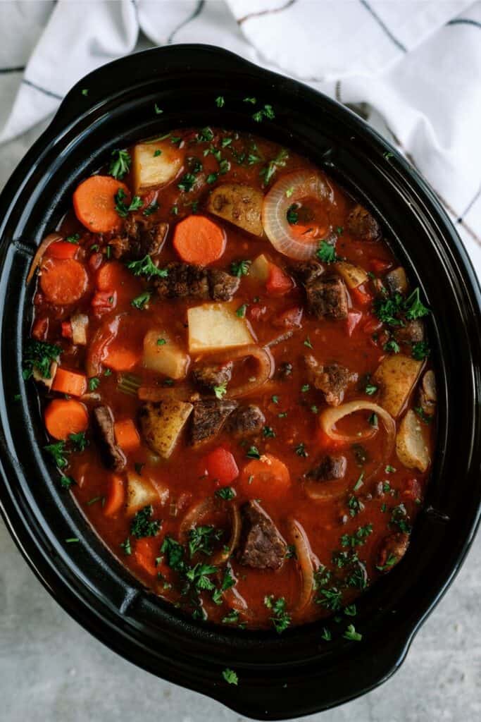 A black pot filled with beef stew, featuring pieces of beef, potatoes, carrots, and onions in a red broth, garnished with chopped parsley on top.