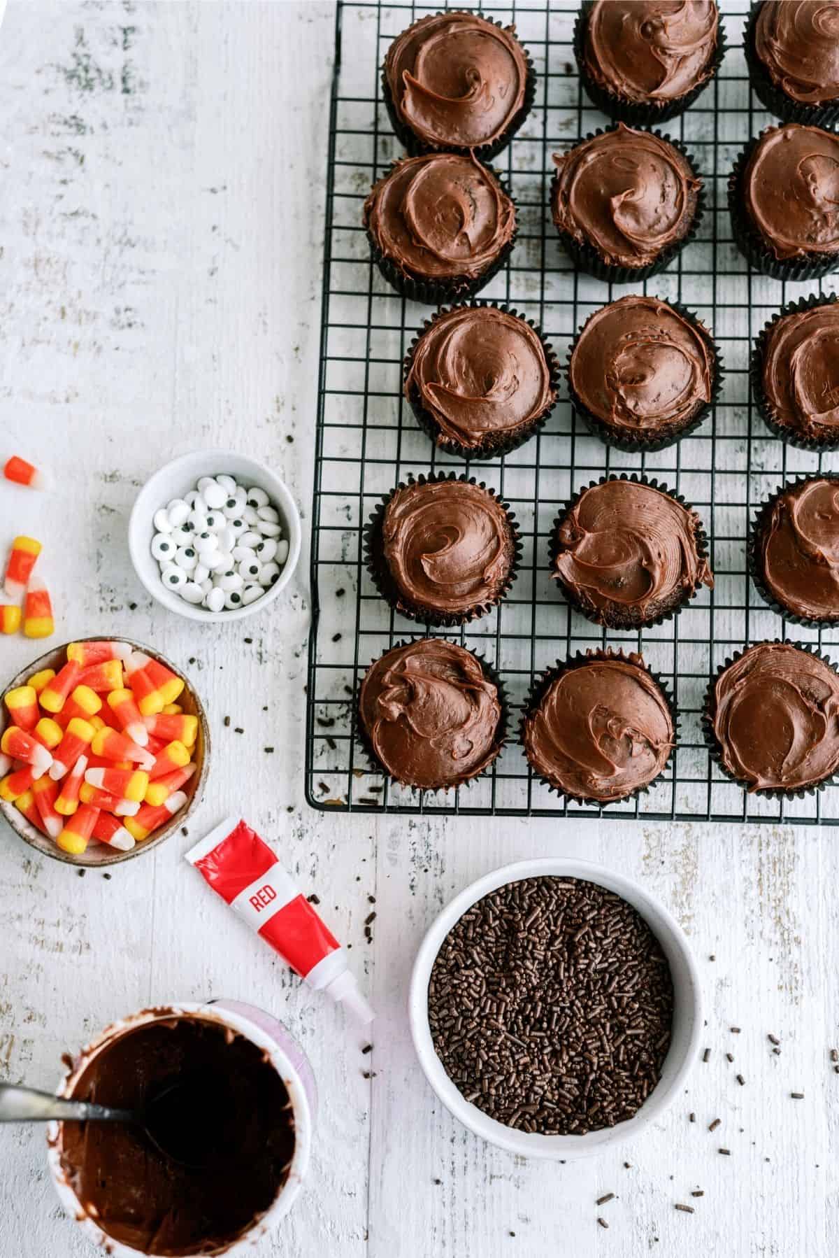 Chocolate frosted cupcakes on a cooling rack with bowls of candy corn, candy eyes, chocolate sprinkles, and a red icing tube on a white wooden surface.