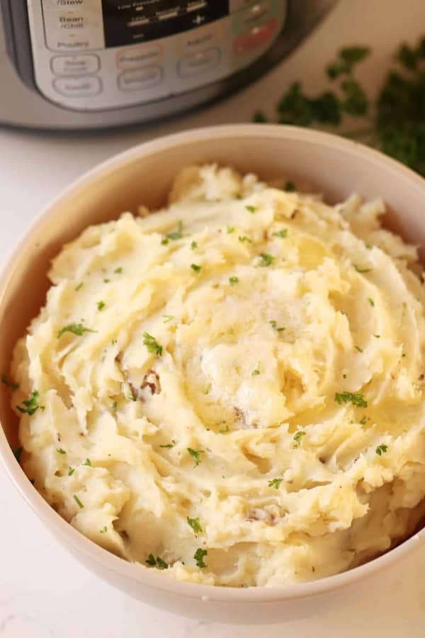 A bowl of mashed potatoes garnished with chopped parsley sits in front of an electric pressure cooker.