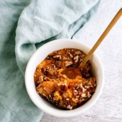 A white ramekin filled with sweet potato casserole topped with chopped pecans, with a gold spoon inside, sits next to a light blue cloth.