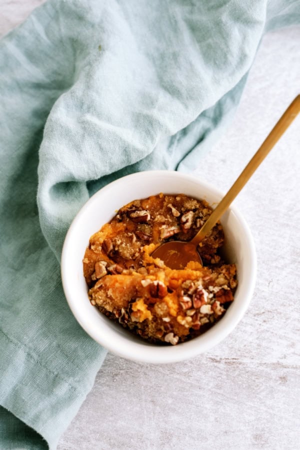 A white ramekin filled with sweet potato casserole topped with chopped pecans, with a gold spoon inside, sits next to a light blue cloth.