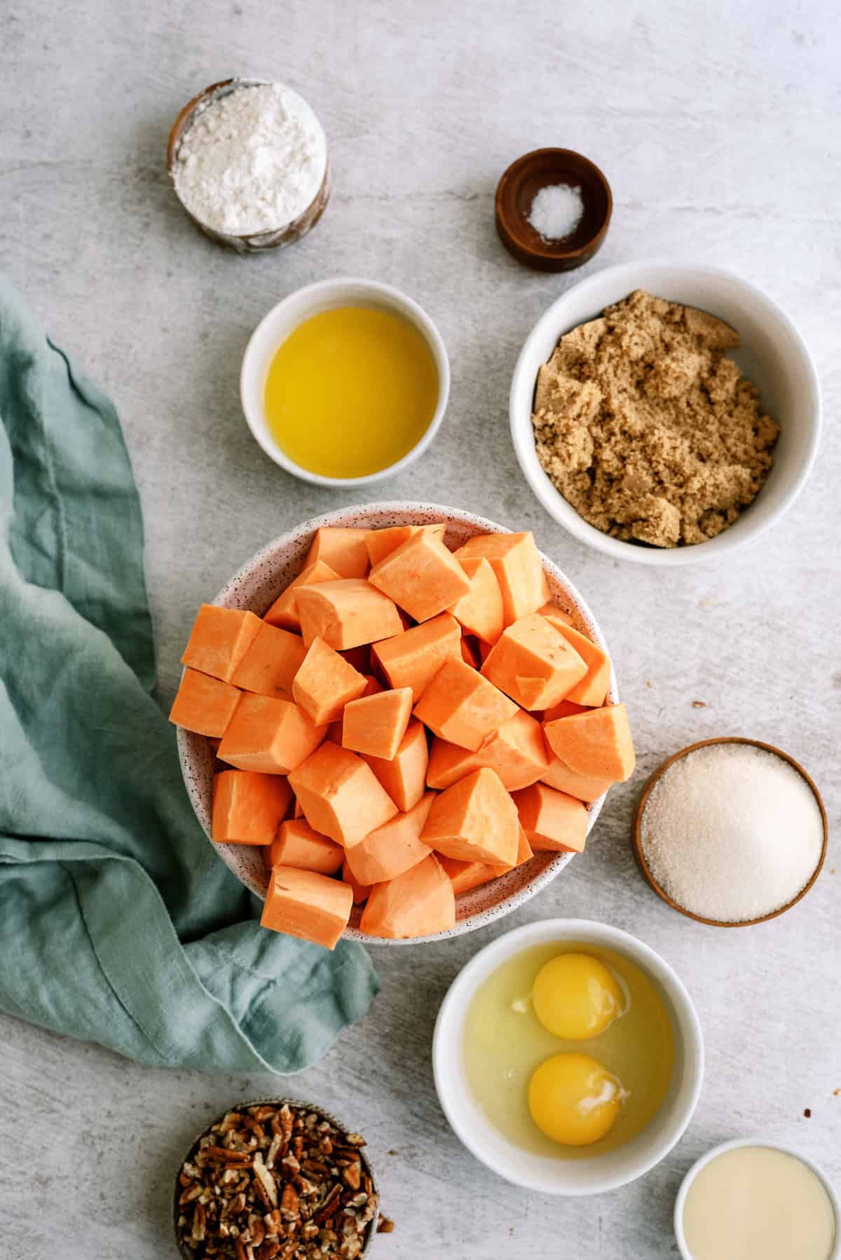 Overhead view of baking ingredients including cubed sweet potatoes, eggs, brown sugar, white sugar, melted butter, flour, salt, and chopped pecans arranged on a light surface.