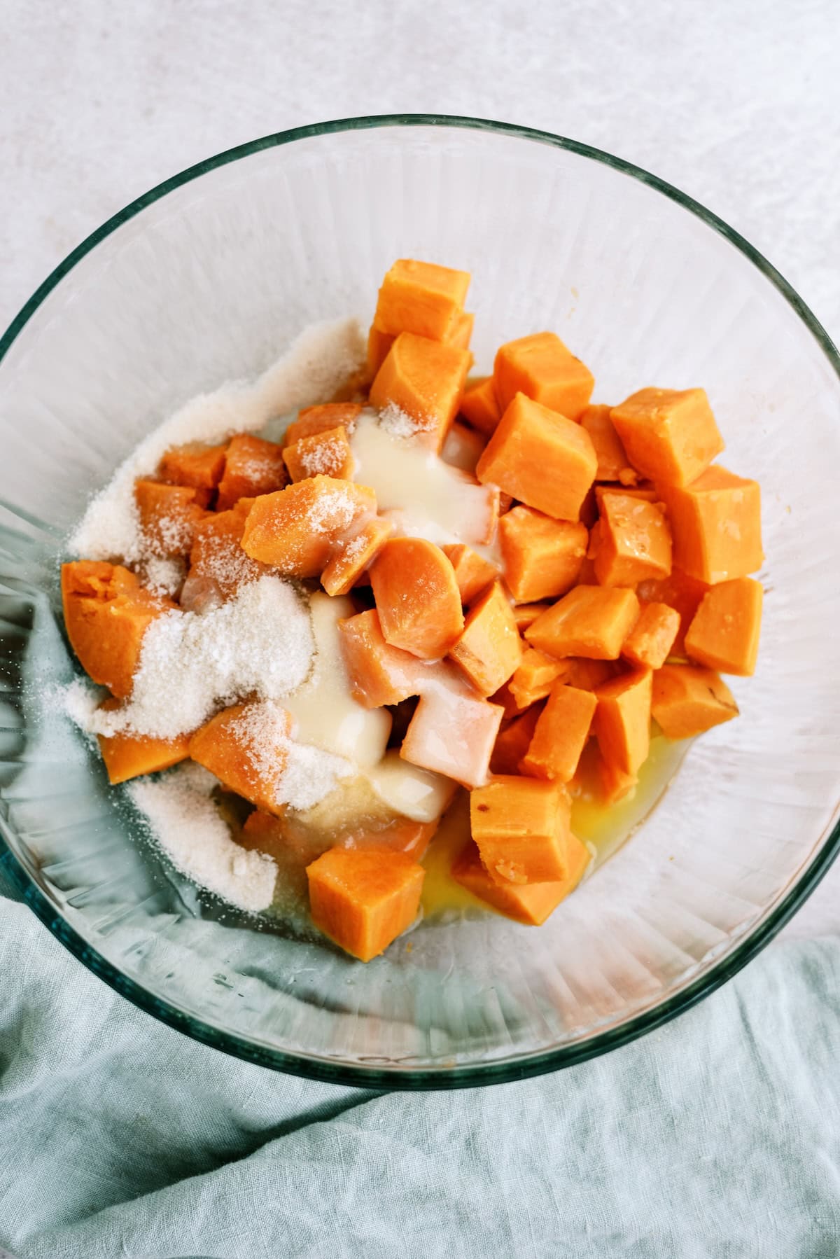 Glass bowl containing cubed sweet potatoes, granulated sugar, melted butter, and milk, all ready to be mixed together.