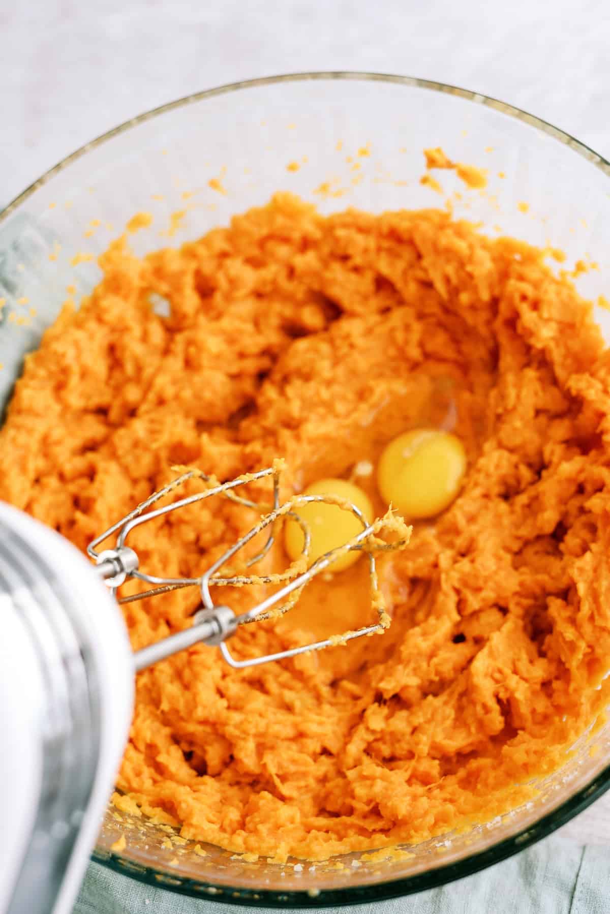 A glass bowl filled with mashed sweet potatoes and two raw eggs, with an electric mixer partially visible.
