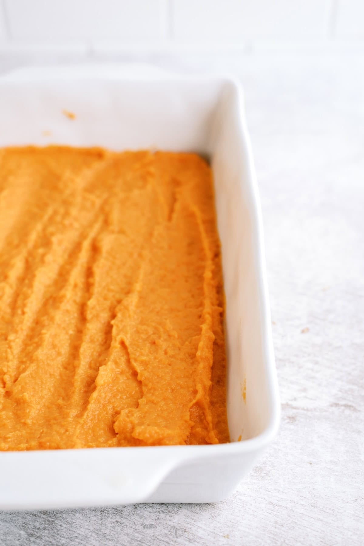 A rectangular white ceramic dish filled with a smooth layer of orange sweet potato mixture, sitting on a light-colored surface.