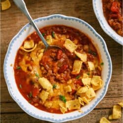 A bowl of chili topped with corn chips and chopped chives sits on a wooden table, with a spoon in the bowl and scattered chips around it.