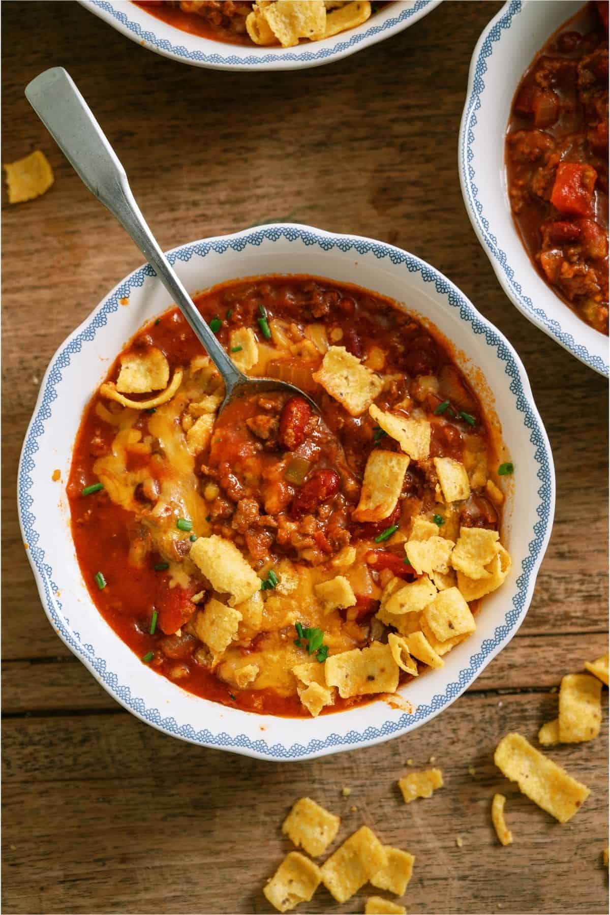 A bowl of chili topped with corn chips and chopped chives sits on a wooden table, with a spoon in the bowl and scattered chips around it.
