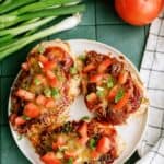 A plate with three baked chicken breasts topped with melted cheese, chopped tomatoes, and sliced green onions, next to fresh tomatoes and green onions on a green tiled surface.