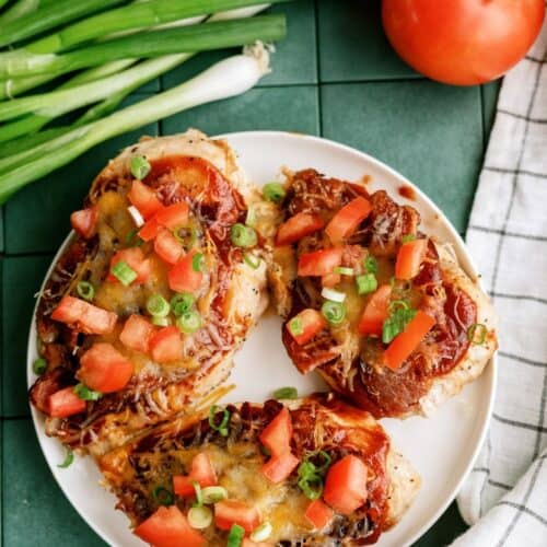 A plate with three baked chicken breasts topped with melted cheese, chopped tomatoes, and sliced green onions, next to fresh tomatoes and green onions on a green tiled surface.