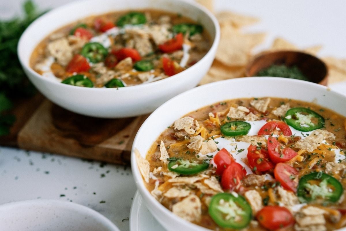 Slow Cooker Nacho Grande Soup in two white bowls topped with tomatoes, tortilla chips and peppers