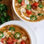 Two bowls of soup topped with sliced jalapeños, cherry tomatoes, crushed tortilla chips, and herbs, set on a wooden and white surface with fresh cilantro nearby.