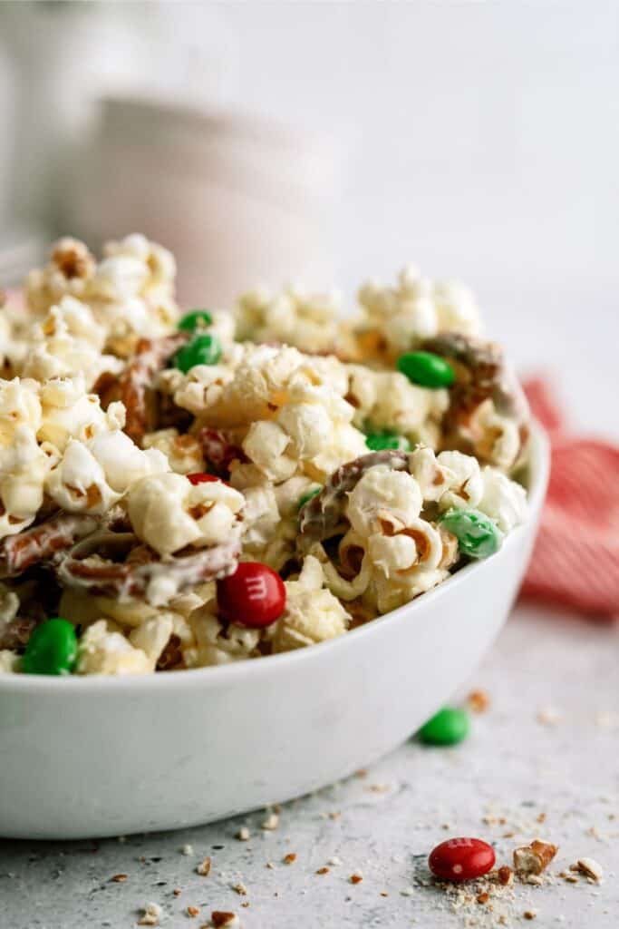 A bowl of popcorn mixed with pretzels and colorful candy-coated chocolates on a light background.