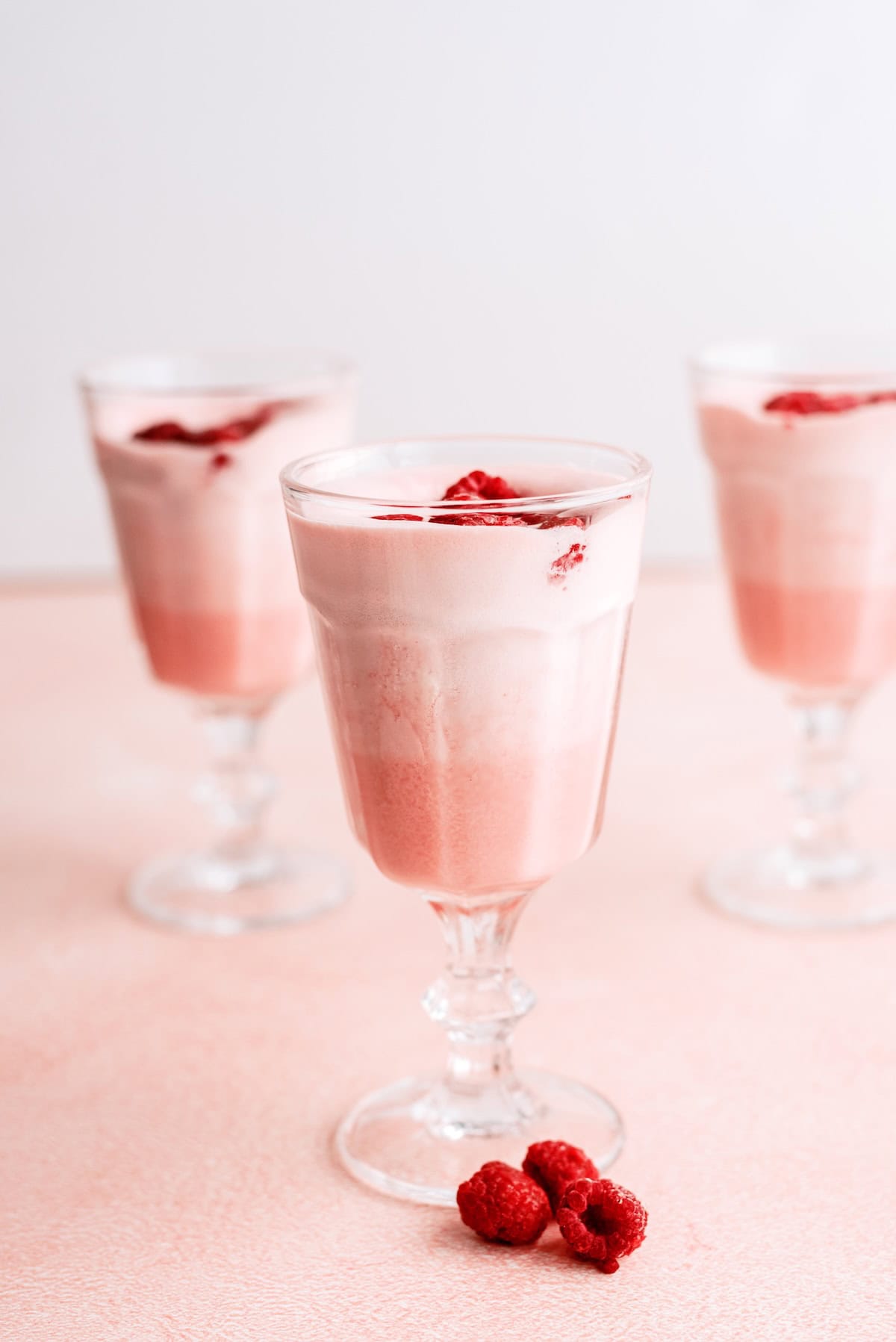 Three glass cups filled with layered pink and white mousse, topped with raspberries, are arranged on a light pink surface with a few raspberries in front.