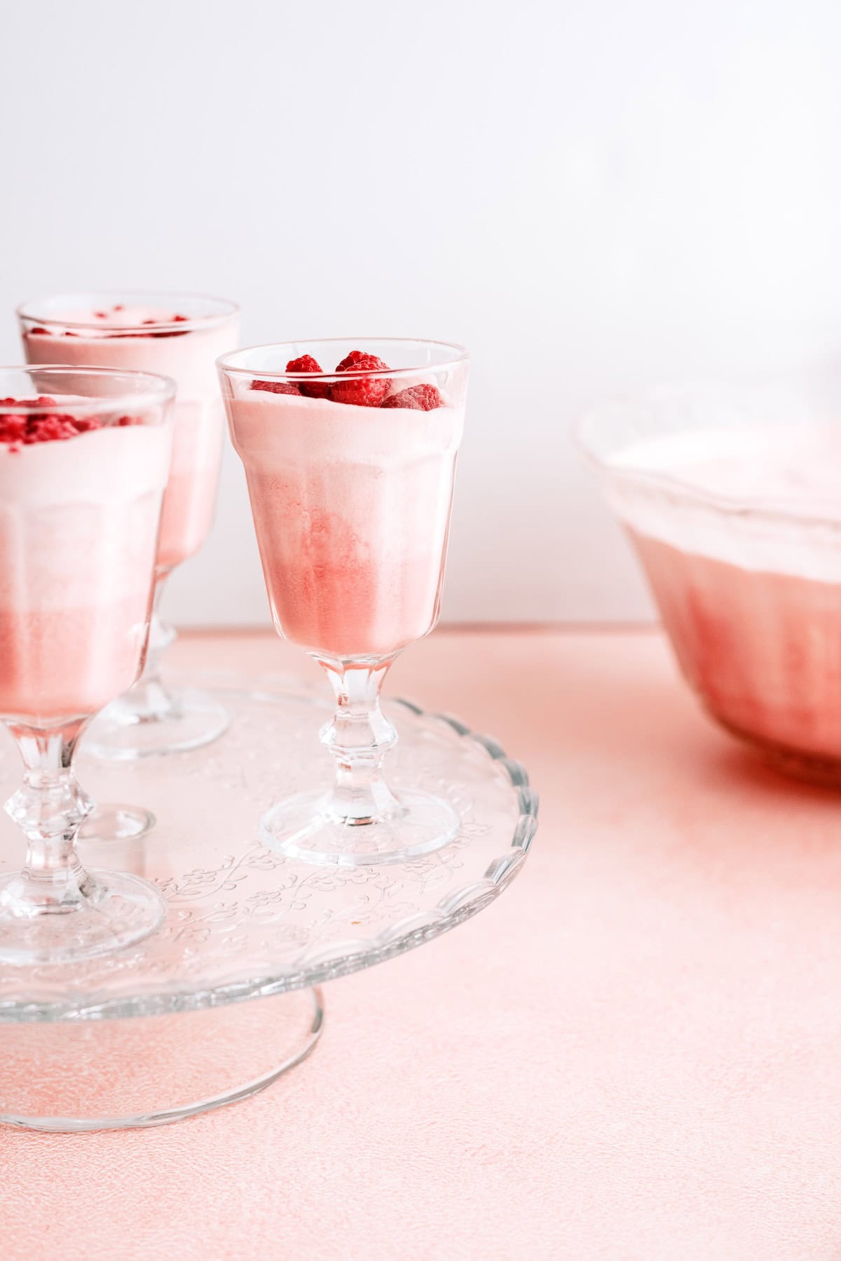 Three glasses of pink mousse topped with strawberries sit on a glass tray, with a large bowl of the same dessert in the background on a light pink surface.
