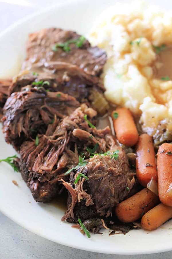Tender beef pot roast with carrots and mashed potatoes on a white plate.