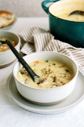A bowl of creamy soup with vegetables and mushrooms, served with a black spoon inside. A striped cloth is in the background.