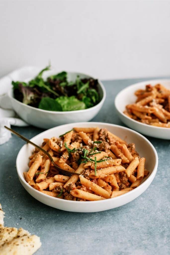 Creamy Sausage and Tomato Pasta in a white bowl with a bowl of spinach in the background.