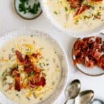 Two bowls of creamy soup topped with shredded cheese, crispy bacon, and chopped chives, with extra bacon and chopped chives on the side, and two spoons nearby.