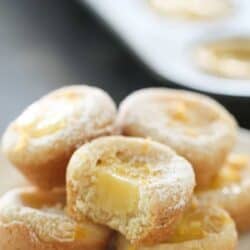 Lemon Bar Cookie Cups stacked on a plate with a muffin tin in the background filled with more Lemon Bar Cookie Cups.