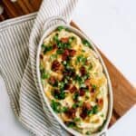 An oval baking dish of mashed potatoes topped with chopped bacon, green onions, and shredded cheese, placed on a striped cloth and wooden board.