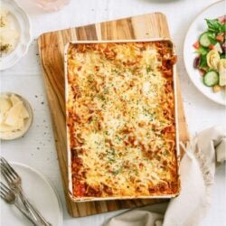 A baked lasagna dish on the table surrounded by cheese, bread and salad.