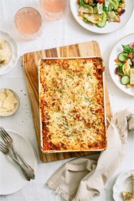 A baked lasagna dish on the table surrounded by cheese, bread and salad.