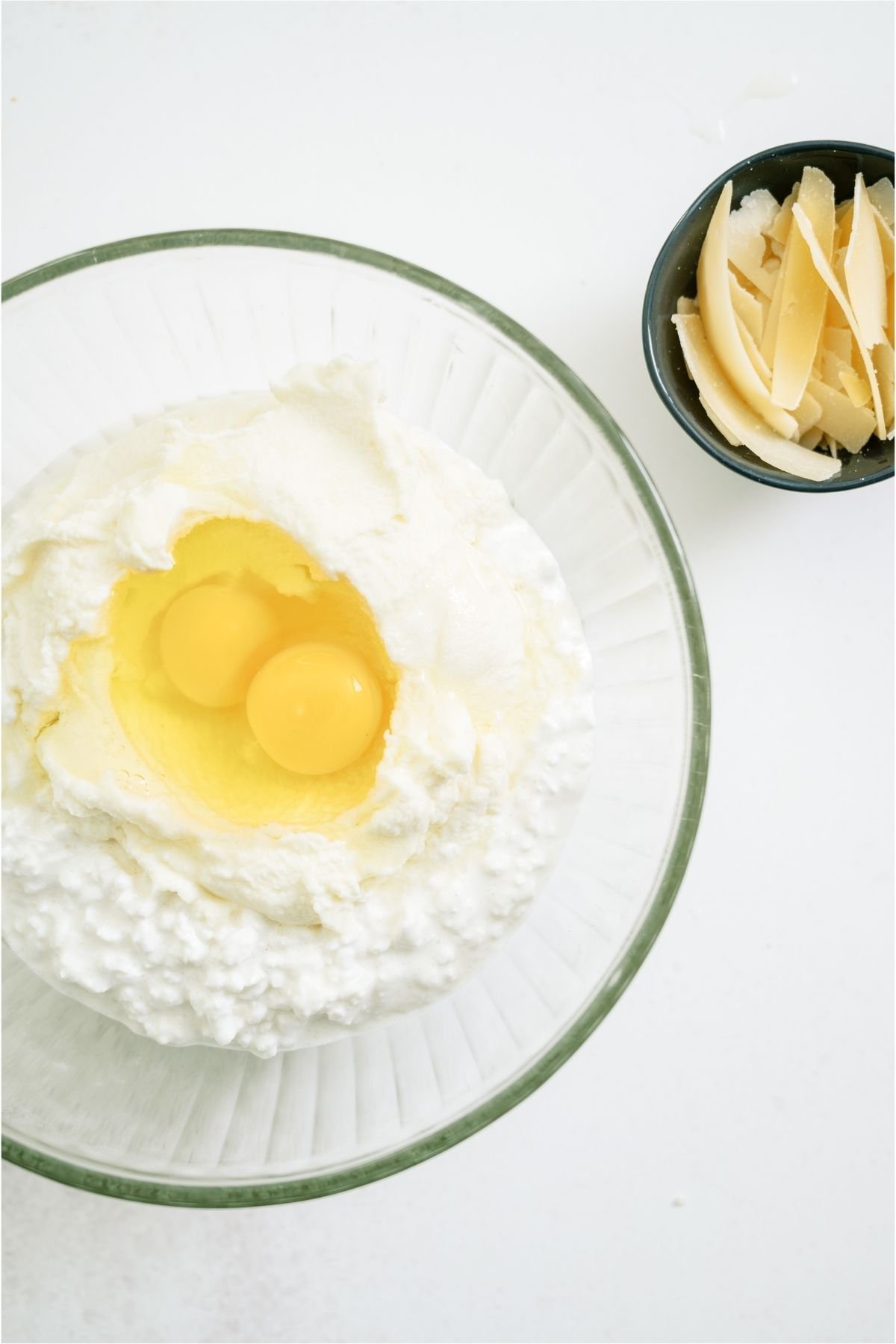 Eggs and cheese in a glass mixing bowl with shredded cheese on the side.