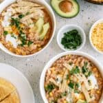 Two bowls of chicken tortilla soup topped with avocado slices, tortilla strips, cilantro, and cream, next to bowls of shredded cheese, chopped herbs, half an avocado, and cornbread.