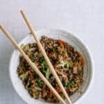 A bowl of ground meat stir-fry with vegetables, garnished with green onions and sesame seeds, with wooden chopsticks resting on top.