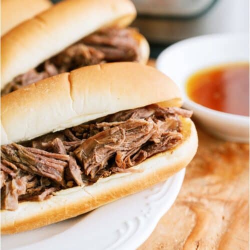 Two Instant Pot French Dip Sandwiches on a white plate with a small bowl of au ju sauce off to the side and an Instant Pot in the background.