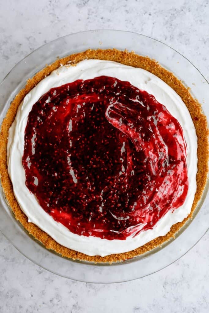 A pie with a graham cracker crust, topped with a layer of white cream and a glossy red berry sauce, on a light-colored surface.