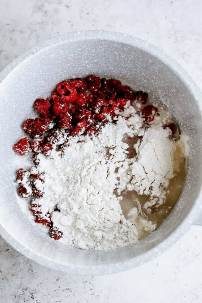 A mixing bowl containing raspberries, flour, and a liquid ingredient on a light-colored surface.