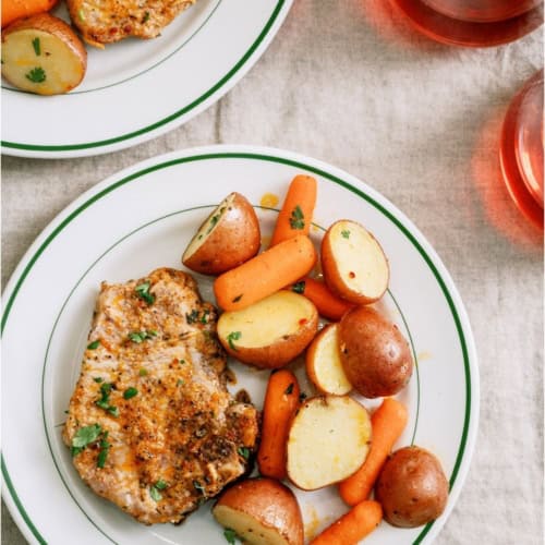Two plates with grilled pork chops, roasted red potatoes, and baby carrots are set on a cream tablecloth, accompanied by two glasses of red beverage.