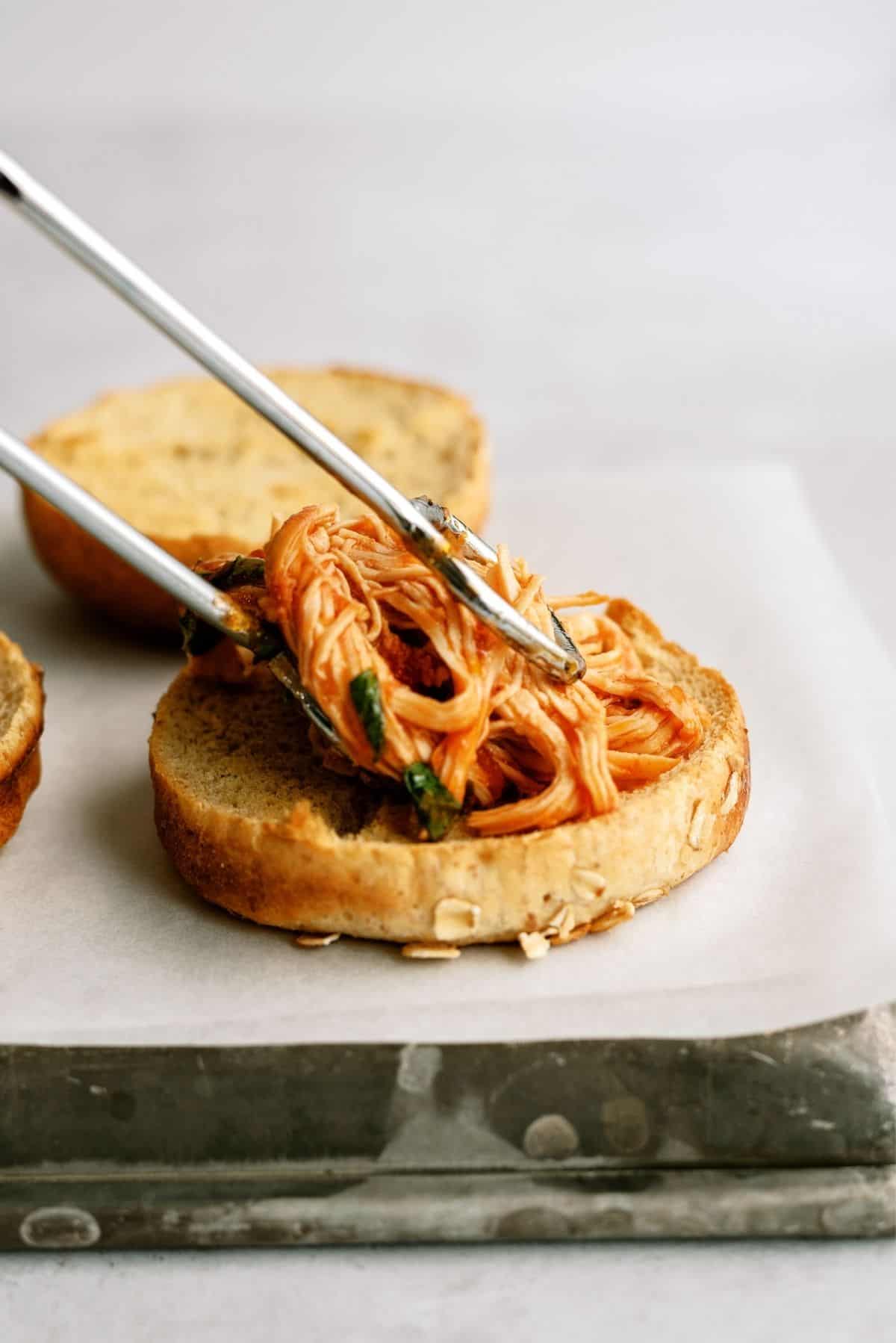 Tongs placing shredded chicken on a bun