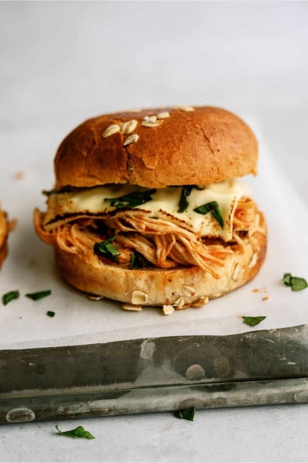 A sandwich with shredded chicken, melted cheese, and greens in a sesame seed bun, placed on white parchment paper.