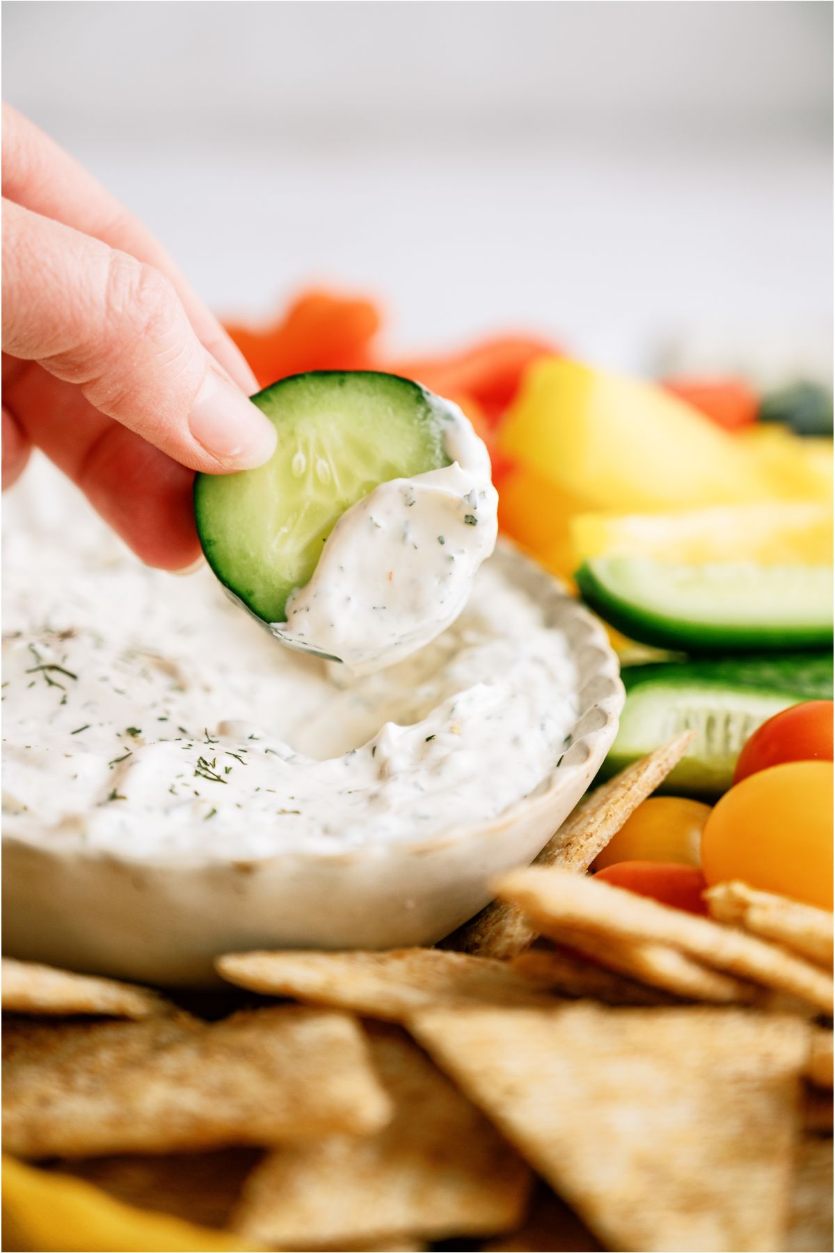 A hand dipping a sliced cucumber into a bowl of Vegetable Dill Dip.