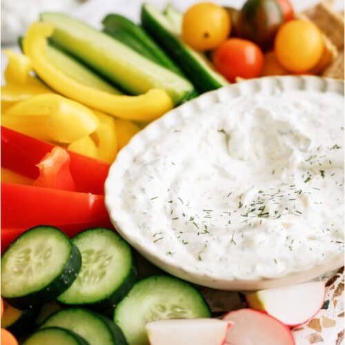 A white bowl of Vegetable Dill Dip on a platter with sliced veggies surrounding it.