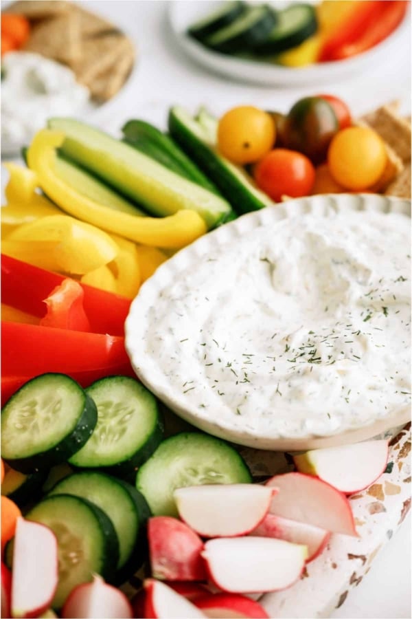 A white bowl of Vegetable Dill Dip on a platter with sliced veggies surrounding it.